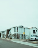 A modern two-story house with a boxy design, featuring large vertical windows and a spiral staircase on the exterior. The building is painted white with a combination of stone elements. The surrounding environment includes a neatly landscaped area with barren trees, and a white SUV is parked on the street. The sky is overcast, creating a muted atmosphere.
