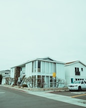 A modern two-story house with a boxy design, featuring large vertical windows and a spiral staircase on the exterior. The building is painted white with a combination of stone elements. The surrounding environment includes a neatly landscaped area with barren trees, and a white SUV is parked on the street. The sky is overcast, creating a muted atmosphere.