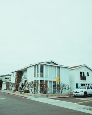 A modern two-story house with a boxy design, featuring large vertical windows and a spiral staircase on the exterior. The building is painted white with a combination of stone elements. The surrounding environment includes a neatly landscaped area with barren trees, and a white SUV is parked on the street. The sky is overcast, creating a muted atmosphere.