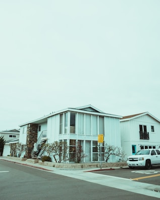 A modern two-story house with a boxy design, featuring large vertical windows and a spiral staircase on the exterior. The building is painted white with a combination of stone elements. The surrounding environment includes a neatly landscaped area with barren trees, and a white SUV is parked on the street. The sky is overcast, creating a muted atmosphere.