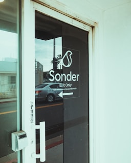 A glass door labeled with 'Sonder Exit Only' in white text with an arrow pointing left. Outside the door, a grey car is parked on the street, and a small building is visible in the background. The handle and lock of the door are also visible.