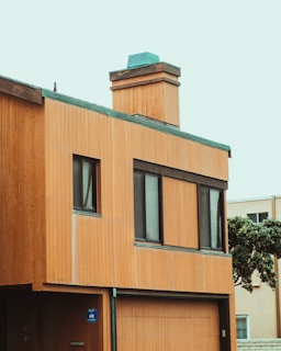 A modern two-story house with wooden paneling and a flat roof. The building has large rectangular windows and a prominent chimney on the top, with green edging contrasting against the wooden textures. A small tree is visible to the side of the house.