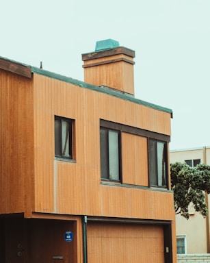 A modern two-story house with wooden paneling and a flat roof. The building has large rectangular windows and a prominent chimney on the top, with green edging contrasting against the wooden textures. A small tree is visible to the side of the house.