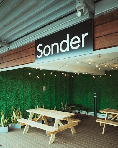 A cozy outdoor seating area features wooden picnic tables placed on a wooden deck. The space is enclosed by lush, green vertical garden walls and decorated with hanging string lights. A sign that reads 'Sonder' is prominently displayed above. A row of potted plants adds to the greenery.