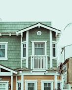 A two-story house features an upper floor with green wooden shingles and white trim, complete with a small balcony. The lower floor is clad in beige siding with several windows. A palm tree is visible to the right, set against a pale sky.