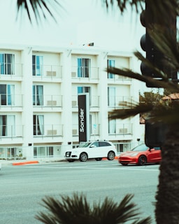 A multi-story building with white walls and balconies, featuring a large vertical sign with the word 'Sonder.' In front of the building, a white SUV and a red sports car are parked on the street. Several palm fronds partially obscure the view, and there is a traffic light pole on the right side.