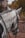 A close-up of the side and back of a white horse's head and neck. The horse has its mane styled in small, neat braids, and is wearing a bridle. The background shows a structure, possibly a stable, with foliage and a wooden fence.