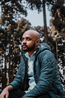 A man with a bald head and beard is wearing a teal puffer jacket and a light blue shirt, sitting in a forest setting surrounded by tall pine trees. He appears thoughtful or contemplative, gazing upward.