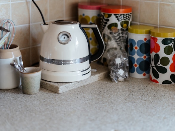 A kitchen countertop features a white electric kettle with a silver band and a temperature gauge. Next to it, there are several colorful patterned storage canisters with retro designs in green, blue, and red. A ceramic container holds kitchen utensils like a whisk and measuring spoons. The background consists of light beige tiled walls.