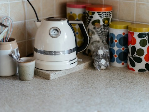 A kitchen countertop features a white electric kettle with a silver band and a temperature gauge. Next to it, there are several colorful patterned storage canisters with retro designs in green, blue, and red. A ceramic container holds kitchen utensils like a whisk and measuring spoons. The background consists of light beige tiled walls.