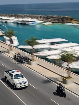 A police pickup truck and a motorcyclist are moving on a road adjacent to a marina with several boats docked. Palm trees line the street, and the ocean is visible in the background.
