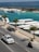 A police pickup truck and a motorcyclist are moving on a road adjacent to a marina with several boats docked. Palm trees line the street, and the ocean is visible in the background.