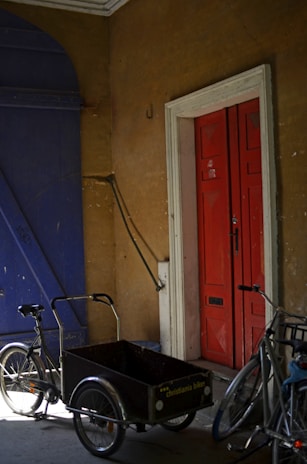 In a dimly lit space, two bicycles are positioned near a robust red door with prominent wooden panels. The bicycles include a classic model and a cargo bike with a labeled compartment. The setting features an exposed wall with a rough texture and a large, dark blue gate, creating a rustic atmosphere with wear and shadow interplay.