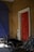 In a dimly lit space, two bicycles are positioned near a robust red door with prominent wooden panels. The bicycles include a classic model and a cargo bike with a labeled compartment. The setting features an exposed wall with a rough texture and a large, dark blue gate, creating a rustic atmosphere with wear and shadow interplay.