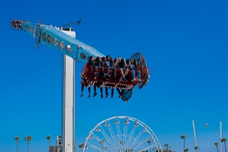 A group of people enjoying a thrilling amusement park ride that swings and spins them high above the ground. The ride is painted bright blue with circular lights underneath it. In the background, a Ferris wheel and several tall palm trees can be seen against a clear blue sky.