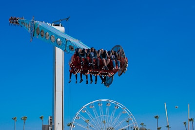 A group of people enjoying a thrilling amusement park ride that swings and spins them high above the ground. The ride is painted bright blue with circular lights underneath it. In the background, a Ferris wheel and several tall palm trees can be seen against a clear blue sky.