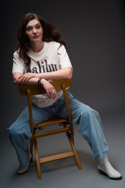 A woman with long, dark hair is seated on a wooden chair, leaning forward with her arms crossed on the back rest. She is wearing a white t-shirt with text that reads 'Malibu', blue jeans, and white ankle boots. The setting is a simple, dark studio background, conveying a relaxed and casual pose.