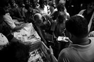 A group of people are gathered closely around a person handing out newspapers. The scene is busy, with everyone focused on receiving the papers. The lighting is dramatic, highlighting the expressions on people's faces.