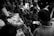 A group of people are gathered closely around a person handing out newspapers. The scene is busy, with everyone focused on receiving the papers. The lighting is dramatic, highlighting the expressions on people's faces.