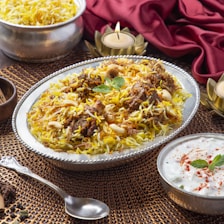 A plate of biryani garnished with mint leaves, cashews, and pieces of meat. The dish is served on an ornate silver platter, surrounded by decorative elements including a red cloth and candle holders. A serving of raita in a small bowl and a silver spoon are placed nearby.