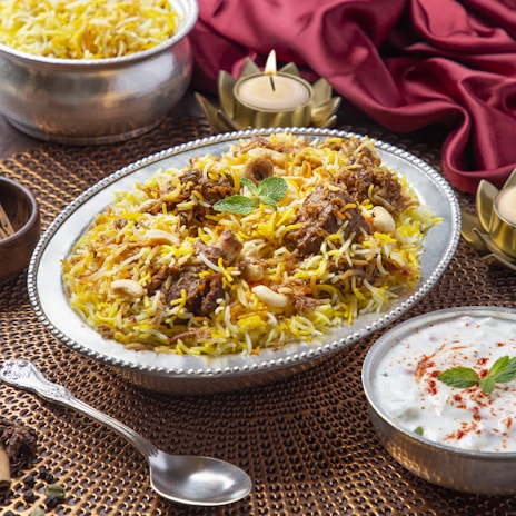 A plate of biryani garnished with mint leaves, cashews, and pieces of meat. The dish is served on an ornate silver platter, surrounded by decorative elements including a red cloth and candle holders. A serving of raita in a small bowl and a silver spoon are placed nearby.