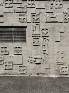 A textured concrete wall with geometric patterns in relief, featuring rectangular and arch-like shapes. There is a grid-patterned window on the left side.