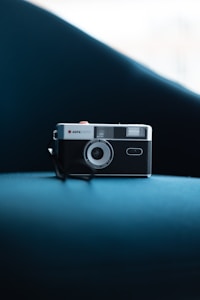 A vintage Agfa camera is placed on a smooth, blue surface with a soft-focus background. The camera's classic design features knobs and a prominent lens, evoking a sense of nostalgia.