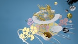 A surreal composition featuring intertwined golden branches, geometric spheres with various textures, and floating white plates. Colorful floral patterns reference delicate organic forms. The background is a pastel blue, providing contrast against the diverse shapes and colors in the scene.