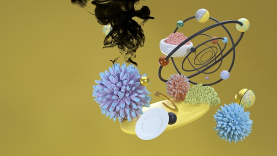 A surreal composition featuring abstract 3D objects including spherical, spiky shapes resembling viruses, a yellow spinning disc, and an orbit-like structure with multiple orbs. Black ink-like fluid is suspended in mid-air above the objects, set against a mustard yellow background.