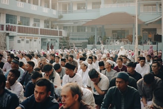 A large gathering of people, mostly men, seated outdoors in what appears to be a courtyard of a building. The attendees are dressed in modest clothing, some wearing religious caps. The atmosphere seems calm and focused, as the participants appear to be engaged in a communal activity or gathering. The building in the background has a pale blue facade and multiple balconies, suggesting it might be an institutional or educational facility.