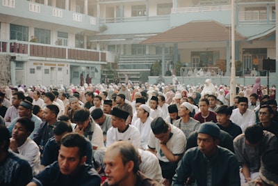 A large gathering of people, mostly men, seated outdoors in what appears to be a courtyard of a building. The attendees are dressed in modest clothing, some wearing religious caps. The atmosphere seems calm and focused, as the participants appear to be engaged in a communal activity or gathering. The building in the background has a pale blue facade and multiple balconies, suggesting it might be an institutional or educational facility.