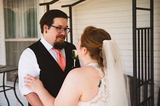A couple stands together, gazing at each other with affection. The man is dressed in a formal outfit with a black vest and peach-colored tie, while the woman is wearing a bridal gown with lace details and a veil. The background features a porch with white siding and a window with curtains.