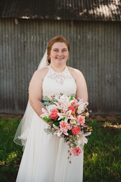A person in a white lace wedding dress holding a bouquet of flowers featuring pink lilies and roses. The background consists of a corrugated metal wall and grass.
