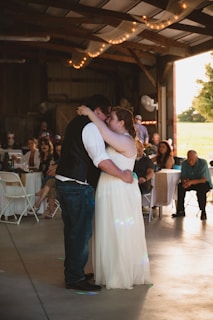 A couple is embracing on a dance floor surrounded by seated guests. The setting appears to be a rustic venue with wooden beams and warm string lights overhead. The bride is wearing a long white gown, and the groom is dressed in dark attire with a white shirt. Guests are casually dressed, observing or enjoying the moment.