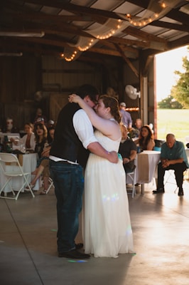 A couple is embracing on a dance floor surrounded by seated guests. The setting appears to be a rustic venue with wooden beams and warm string lights overhead. The bride is wearing a long white gown, and the groom is dressed in dark attire with a white shirt. Guests are casually dressed, observing or enjoying the moment.