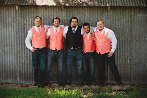 Five men are standing casually with their arms around each other in front of a corrugated metal wall. Four of them are wearing coral-colored vests with white shirts and ties, while one is in a black vest with a blue tie. They stand on a grassy patch with rocks at their feet, displaying a relaxed and cheerful demeanor.