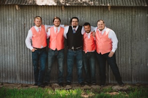 Five men are standing casually with their arms around each other in front of a corrugated metal wall. Four of them are wearing coral-colored vests with white shirts and ties, while one is in a black vest with a blue tie. They stand on a grassy patch with rocks at their feet, displaying a relaxed and cheerful demeanor.