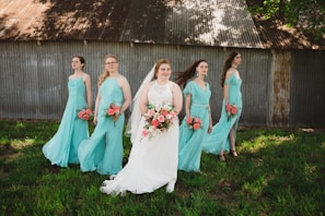 A bride in a white gown stands in the center, holding a bouquet of pink and white flowers. She is surrounded by four bridesmaids wearing matching turquoise dresses, each holding similar bouquets. They are standing on a grassy area with a rustic, corrugated metal building in the background. Shadows from trees are visible on the building.