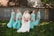 A bride in a white gown stands in the center, holding a bouquet of pink and white flowers. She is surrounded by four bridesmaids wearing matching turquoise dresses, each holding similar bouquets. They are standing on a grassy area with a rustic, corrugated metal building in the background. Shadows from trees are visible on the building.