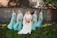 A bride in a white gown stands in the center, holding a bouquet of pink and white flowers. She is surrounded by four bridesmaids wearing matching turquoise dresses, each holding similar bouquets. They are standing on a grassy area with a rustic, corrugated metal building in the background. Shadows from trees are visible on the building.