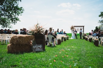 Outdoor wedding ceremony setup on a grassy field with hay bales, decorative flowers, and a wooden arch. Guests are seated on white chairs, and a couple is standing at the altar with an officiant.
