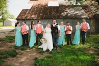 A group of people dressed in formal attire, including a bride with a white gown holding a bouquet, and several bridesmaids and groomsmen wearing matching pastel turquoise and coral outfits. They are standing on grass in front of a rustic barn with a corrugated metal roof.