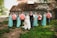 A group of people dressed in formal attire, including a bride with a white gown holding a bouquet, and several bridesmaids and groomsmen wearing matching pastel turquoise and coral outfits. They are standing on grass in front of a rustic barn with a corrugated metal roof.
