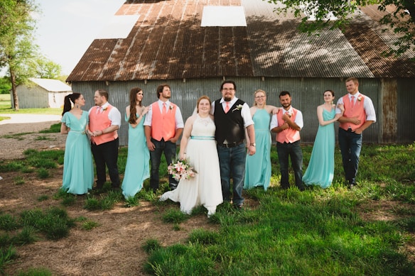 A group of people dressed in formal attire, including a bride with a white gown holding a bouquet, and several bridesmaids and groomsmen wearing matching pastel turquoise and coral outfits. They are standing on grass in front of a rustic barn with a corrugated metal roof.