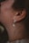 A close-up of a woman's ear adorned with an elegant earring. The earring features a small, clear, teardrop-shaped crystal dangling from a gold setting with smaller crystals and a pearl. The woman's skin has a natural tone, and her hair is brown and loosely tied back.