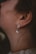A close-up of a woman's ear adorned with an elegant earring. The earring features a small, clear, teardrop-shaped crystal dangling from a gold setting with smaller crystals and a pearl. The woman's skin has a natural tone, and her hair is brown and loosely tied back.