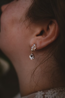 A close-up of a woman's ear adorned with an elegant earring. The earring features a small, clear, teardrop-shaped crystal dangling from a gold setting with smaller crystals and a pearl. The woman's skin has a natural tone, and her hair is brown and loosely tied back.