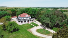 A large, isolated house with a red roof is surrounded by lush green trees and open fields under a clear blue sky. A white gravel driveway leads to the front of the house, which has a wraparound porch.