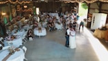 A rustic venue is decorated for a wedding reception featuring round tables with white tablecloths, floral centerpieces, and guests seated around them. A couple is dancing in the center under soft lighting. String lights and draped fabric add to the cozy atmosphere. On the sides, people are observing the dance, and there's a vintage ambiance with wooden walls and soft green accents.