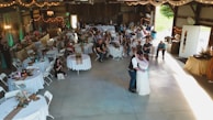 A rustic venue is decorated for a wedding reception featuring round tables with white tablecloths, floral centerpieces, and guests seated around them. A couple is dancing in the center under soft lighting. String lights and draped fabric add to the cozy atmosphere. On the sides, people are observing the dance, and there's a vintage ambiance with wooden walls and soft green accents.